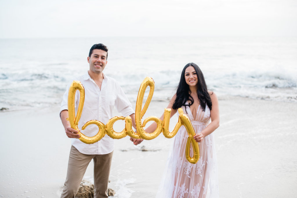couple standing in front of the ocean holding a gold balloon that says baby